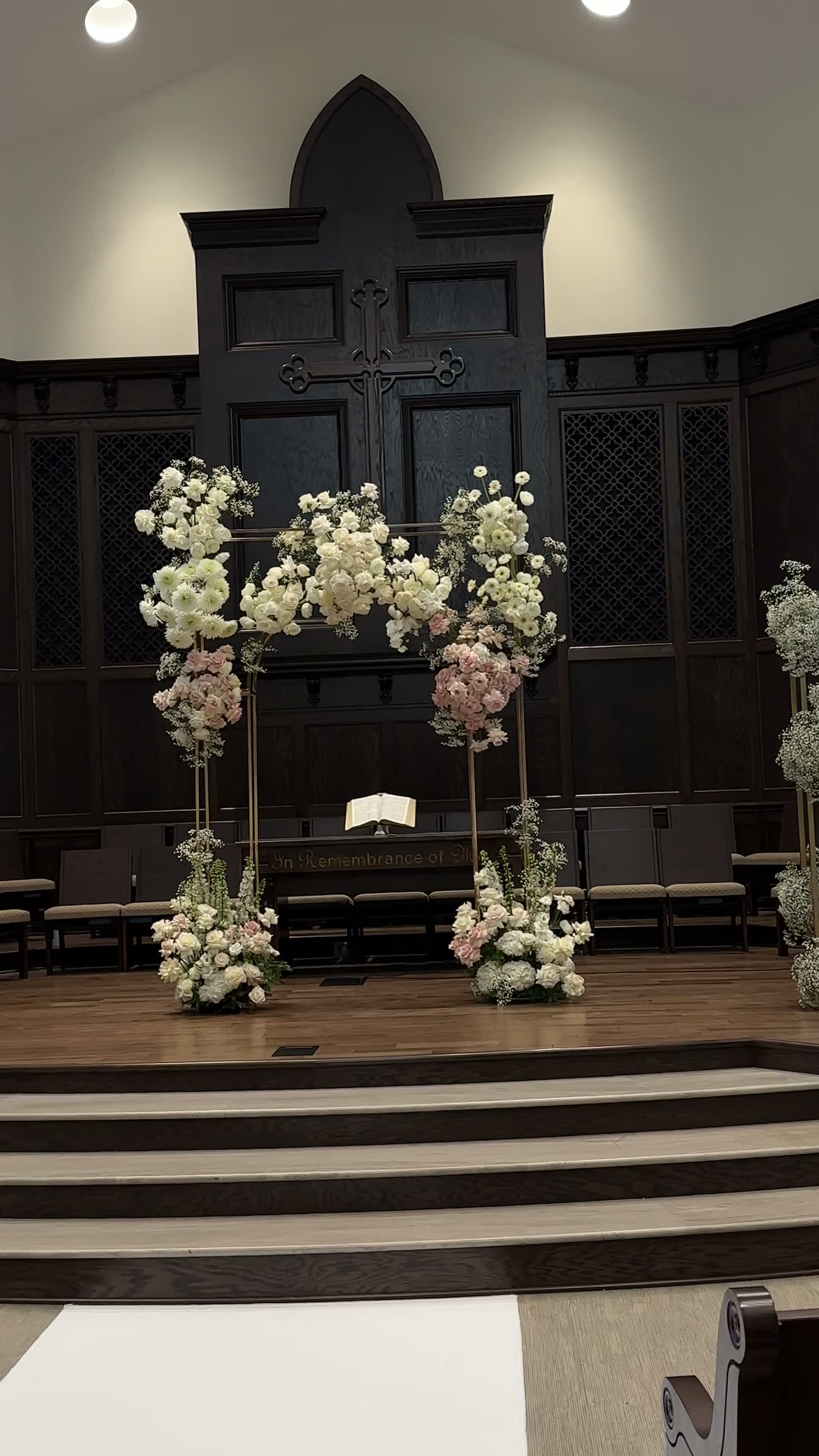 A wedding ceremony floral installation of white roses, blush peonies and baby's breath on gold frames, in a dark-paneled chapel